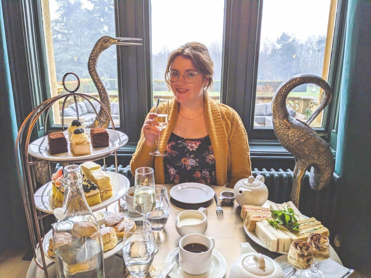 A woman in glasses and a floral dress smiles whilst holding a glass of champagne at a table set for afternoon tea, with cakes, sandwiches, and tea. Two decorative bird sculptures are on either side. Large windows show a garden view.