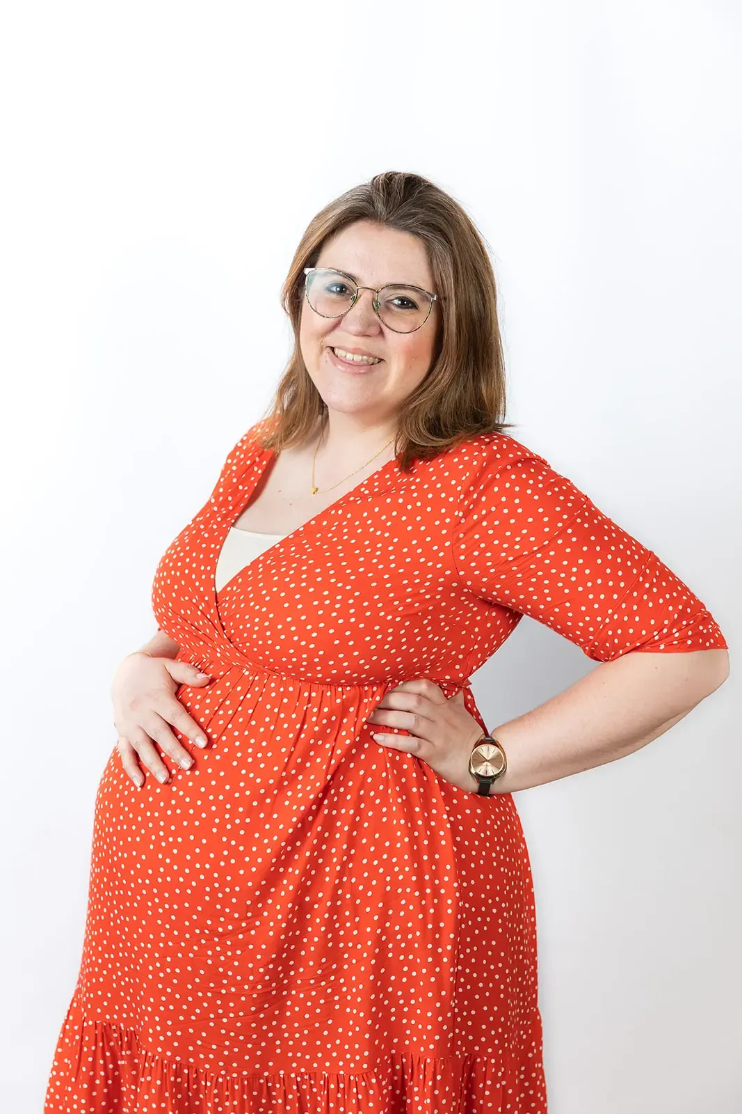 A smiling pregnant virtual assistant with brown hair and glasses wears a red dress with white polka dots. She stands in front of a plain white background, one hand on her bump and the other on her hip.
