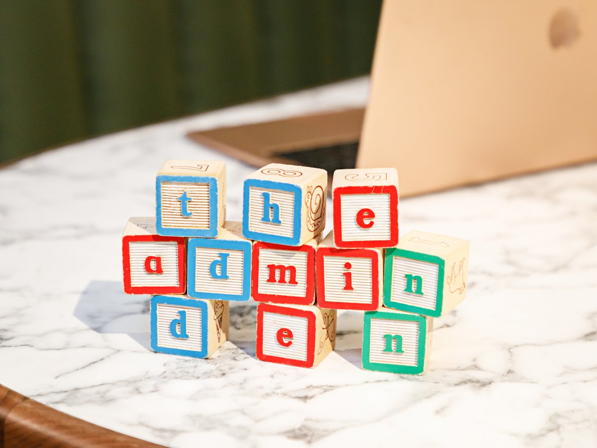 Wooden alphabet blocks spelling out 'the admin den' stacked on a marble table in front of a laptop.