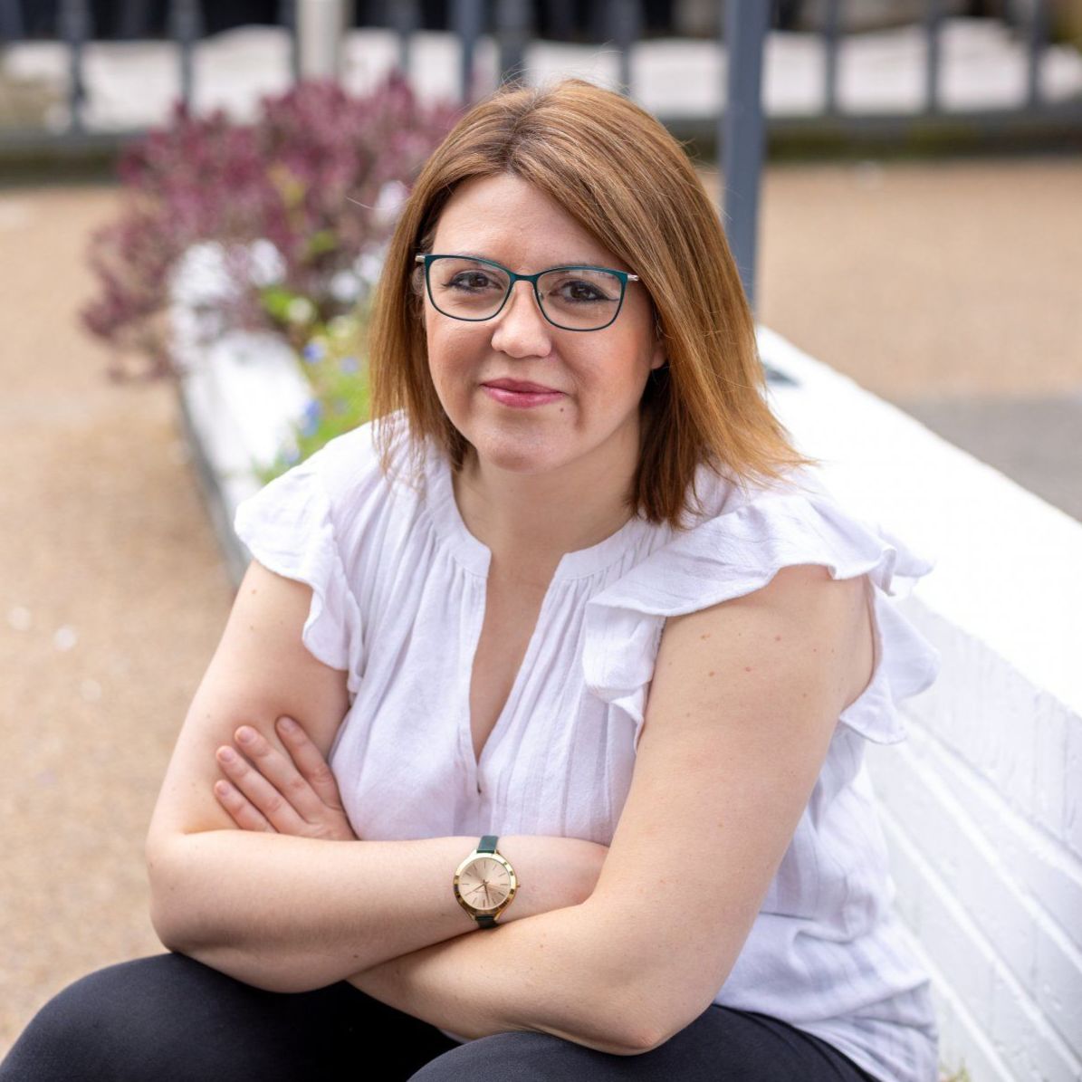 Alicia, Virtual Assistant, sat on a wall with her arms crossed leaning on her knees, she is smiling welcomingly. Alicia is wearing a white frilled sleeveless shirt, black jeans and glasses.