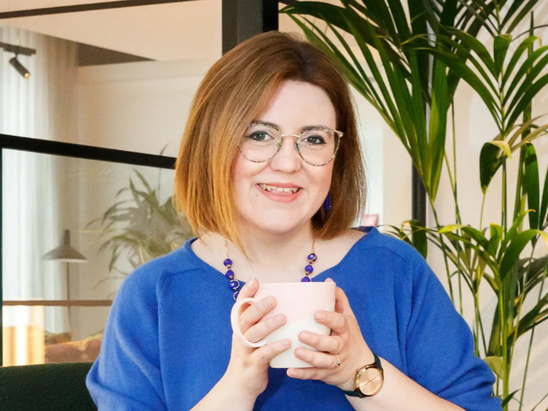 A woman with glasses sits on a green chair in a modern office, holding a white mug and smiling at the camera.