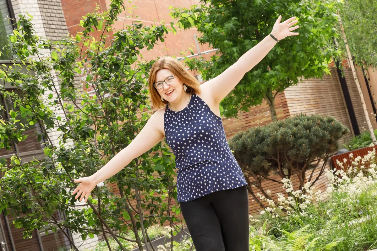 A virtual assistant with shoulder-length hair and glasses stands outdoors in a garden, smiling with arms outstretched. She wears a sleeveless, navy blue polka dot top and black trousers. Greenery and a brick building are in the background.